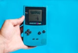 A hand holds a teal Game Boy Color against a matching blue background. The screen displays the title of a Pokémon game. Visible buttons include a directional pad and the A, B, Select, and Start buttons.