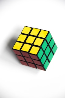 A Rubik's Cube with visible sides showing yellow, green, and red squares on a white background. The cube is rotated, displaying three sides, and appears to be in a solved state.