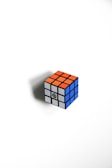 A classic Rubik's Cube with a mix of orange, white, and blue squares is positioned on a white background. The cube casts a subtle shadow to the left, suggesting a light source coming from the right side.