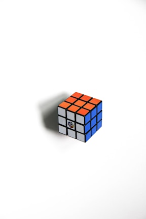 A classic Rubik's Cube with a mix of orange, white, and blue squares is positioned on a white background. The cube casts a subtle shadow to the left, suggesting a light source coming from the right side.