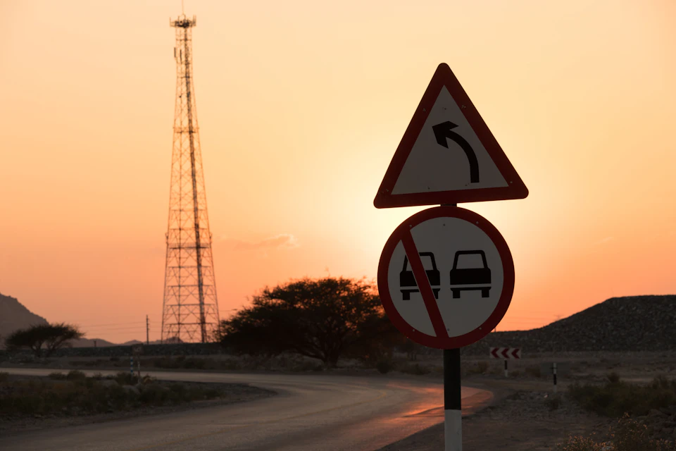 The image depicts a rural landscape at sunset, with a tall tower standing in the background and a road winding through it. On the right side of the road, there is a triangular traffic sign displaying an arrow pointing to the left, indicating that vehicles are allowed to turn left on this particular stretch of road. The sky above is painted in warm hues of orange and yellow, creating a serene atmosphere as the sun sets over the horizon.