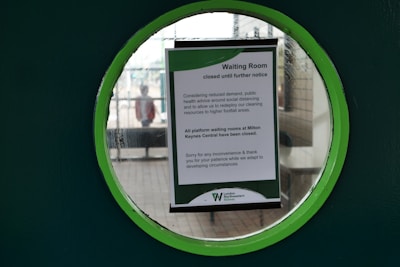 A notice displayed on a circular window announces the closure of a waiting room until further notice. The window is green, and outside, a blurred figure of a person can be seen near a bench on a platform. The notice cites social distancing and cleaning resource allocation as reasons for the closure.