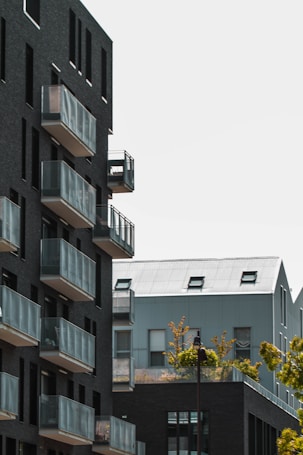 The image depicts modern apartment buildings with dark brick facades and large glass balconies. In the background, there is a metal-roofed building partially visible, with small windows and a couple of skylights. Trees with green foliage are visible in front of the buildings, adding a touch of nature to the urban landscape.