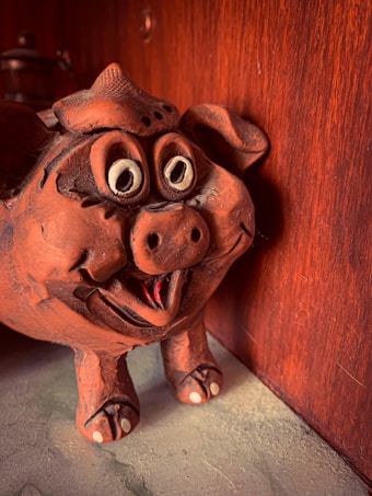 A clay sculpture of a pig with exaggerated facial features, including large eyes and a wide-open mouth. The texture of the clay is rough, with a deep reddish-brown color. It appears to be positioned against a wooden surface which enhances its rustic appearance.
