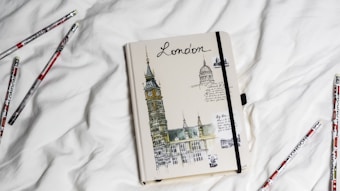 A notebook with a cover featuring sketches of iconic London landmarks including the Big Ben and St. Paul's Cathedral, resting on a white surface. Surrounding the notebook are several pencils with the word 'London' and graphic designs printed on them.