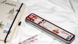 A notebook and pencil case with London-themed designs are placed on a white surface. The notebook features sketches and text related to London, with a black elastic band around it. The pencil case is metallic with red, white, and black colors, depicting icons such as a double-decker bus and a soldier. Two pencils with similar London motifs are nearby.