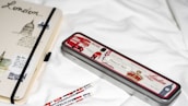 A notebook and pencil case with London-themed designs are placed on a white surface. The notebook features sketches and text related to London, with a black elastic band around it. The pencil case is metallic with red, white, and black colors, depicting icons such as a double-decker bus and a soldier. Two pencils with similar London motifs are nearby.