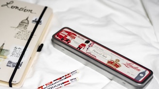 A notebook and pencil case with London-themed designs are placed on a white surface. The notebook features sketches and text related to London, with a black elastic band around it. The pencil case is metallic with red, white, and black colors, depicting icons such as a double-decker bus and a soldier. Two pencils with similar London motifs are nearby.