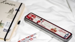 A notebook and pencil case with London-themed designs are placed on a white surface. The notebook features sketches and text related to London, with a black elastic band around it. The pencil case is metallic with red, white, and black colors, depicting icons such as a double-decker bus and a soldier. Two pencils with similar London motifs are nearby.
