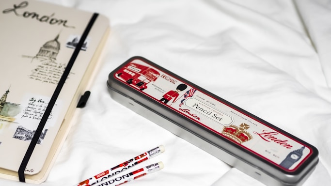 A notebook and pencil case with London-themed designs are placed on a white surface. The notebook features sketches and text related to London, with a black elastic band around it. The pencil case is metallic with red, white, and black colors, depicting icons such as a double-decker bus and a soldier. Two pencils with similar London motifs are nearby.