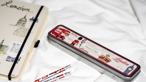 A notebook and pencil case with London-themed designs are placed on a white surface. The notebook features sketches and text related to London, with a black elastic band around it. The pencil case is metallic with red, white, and black colors, depicting icons such as a double-decker bus and a soldier. Two pencils with similar London motifs are nearby.