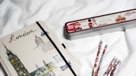 A notebook with illustrations of London landmarks and the word 'London' written on the cover lies on a white surface next to a tin pencil case decorated with London-themed graphics and several pencils. The pencil case has images of a double-decker bus, a traditional guard, and British symbols.