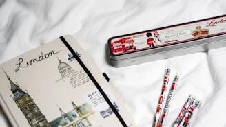 A notebook with illustrations of London landmarks and the word 'London' written on the cover lies on a white surface next to a tin pencil case decorated with London-themed graphics and several pencils. The pencil case has images of a double-decker bus, a traditional guard, and British symbols.
