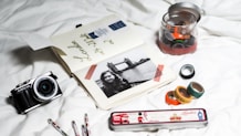 A collection of travel-related items arranged on a white surface, including an open journal with travel notes and a black-and-white photo of a woman with Tower Bridge in the background. There is a vintage-style camera on the left and a tin pencil case labeled 'London' with British-themed images. Several washi tapes in a variety of colors and patterns are scattered, some inside a jar and others outside.