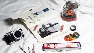 A collection of travel-related items arranged on a white surface, including an open journal with travel notes and a black-and-white photo of a woman with Tower Bridge in the background. There is a vintage-style camera on the left and a tin pencil case labeled 'London' with British-themed images. Several washi tapes in a variety of colors and patterns are scattered, some inside a jar and others outside.