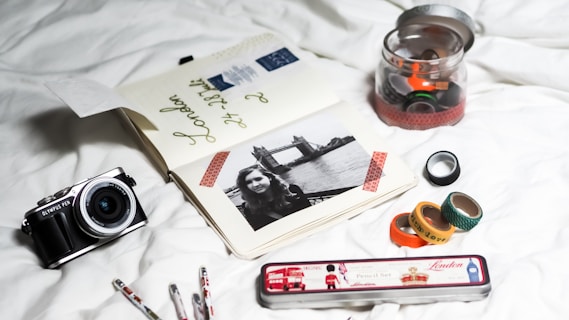 A collection of travel-related items arranged on a white surface, including an open journal with travel notes and a black-and-white photo of a woman with Tower Bridge in the background. There is a vintage-style camera on the left and a tin pencil case labeled 'London' with British-themed images. Several washi tapes in a variety of colors and patterns are scattered, some inside a jar and others outside.