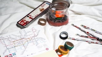 A collection of items is laid out on a white surface, including a map of the London Tube system, rolls of decorative tape, a clear jar with colored tape inside, and several pencils with London-themed designs. The metal tin in the background has illustrations of landmarks or items associated with London.