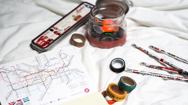 A collection of items is laid out on a white surface, including a map of the London Tube system, rolls of decorative tape, a clear jar with colored tape inside, and several pencils with London-themed designs. The metal tin in the background has illustrations of landmarks or items associated with London.