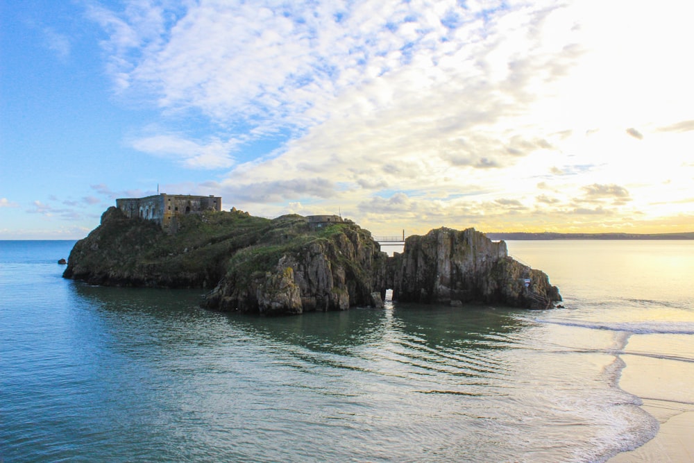 brown concrete building on green rock formation beside sea under blue and white cloudy sky during