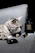 A grey tabby cat is resting on a plush grey couch. In front of the cat are three Chanel products: a perfume bottle, a lipstick, and a compact case. The setting appears to be stylish and luxurious.