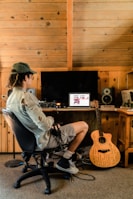 A person sits in a chair facing a computer on a wooden desk in a rustic room with wooden paneling. The computer screen displays music software, and various audio equipment surrounds it. A guitar with writings on its body leans against the desk.