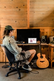 A person sits in a chair facing a computer on a wooden desk in a rustic room with wooden paneling. The computer screen displays music software, and various audio equipment surrounds it. A guitar with writings on its body leans against the desk.