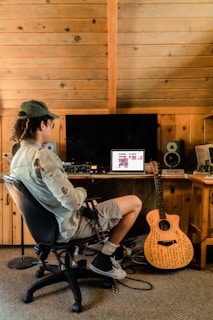 A person sits in a chair facing a computer on a wooden desk in a rustic room with wooden paneling. The computer screen displays music software, and various audio equipment surrounds it. A guitar with writings on its body leans against the desk.