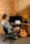 A person sits in a chair facing a computer on a wooden desk in a rustic room with wooden paneling. The computer screen displays music software, and various audio equipment surrounds it. A guitar with writings on its body leans against the desk.