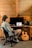 A person sits in a chair facing a computer on a wooden desk in a rustic room with wooden paneling. The computer screen displays music software, and various audio equipment surrounds it. A guitar with writings on its body leans against the desk.