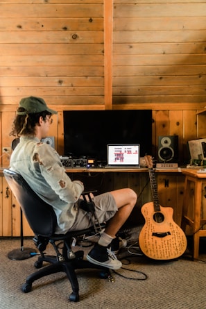 A person sits in a chair facing a computer on a wooden desk in a rustic room with wooden paneling. The computer screen displays music software, and various audio equipment surrounds it. A guitar with writings on its body leans against the desk.