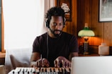 A man with headphones on is playing a keyboard in a cozy, wooden-paneled room. A green lamp emits a warm glow on the side, and there is a framed picture and a small decorative vase behind him. Natural light comes through a window with sheer curtains.
