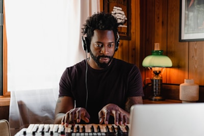 A man with headphones on is playing a keyboard in a cozy, wooden-paneled room. A green lamp emits a warm glow on the side, and there is a framed picture and a small decorative vase behind him. Natural light comes through a window with sheer curtains.