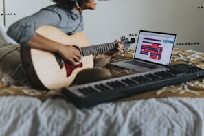 A person sits on a bed playing an acoustic guitar, with a keyboard and a laptop next to them. The laptop screen displays music creation software. The room has a cozy, relaxed atmosphere.