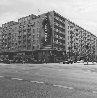 A large urban apartment building with multiple stories and a significant banner displaying 'THE NATIONAL' along one side. Below the building, there are several shops and a KFC restaurant. Cars are parked and moving along the street, with a few pedestrians visible on the sidewalk.