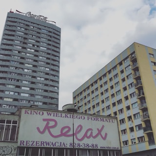 Two tall buildings stand against a cloudy sky, with a large sign advertising a cinema named 'Relax' in the foreground. The building on the left is adorned with a Coca-Cola sign on top, while the other is a residential structure featuring balconies. The sign below contains text in Polish and displays contact information.