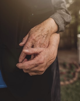 A close-up of elderly hands clasped together, conveying a sense of age and experience. The image is softly lit, highlighting the texture of the skin and the gentle grip.