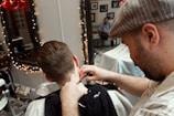 A barber is seen trimming a client's hair using clippers. The focus is on the back of the client's head and the barber's attentive expression as he works. The setting includes a well-decorated barbershop with a mirror adorned with string lights and various framed pictures on the wall. Another person is visible in the background, sitting and observing the scene.