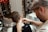 A barber is seen trimming a client's hair using clippers. The focus is on the back of the client's head and the barber's attentive expression as he works. The setting includes a well-decorated barbershop with a mirror adorned with string lights and various framed pictures on the wall. Another person is visible in the background, sitting and observing the scene.