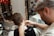 A barber is seen trimming a client's hair using clippers. The focus is on the back of the client's head and the barber's attentive expression as he works. The setting includes a well-decorated barbershop with a mirror adorned with string lights and various framed pictures on the wall. Another person is visible in the background, sitting and observing the scene.