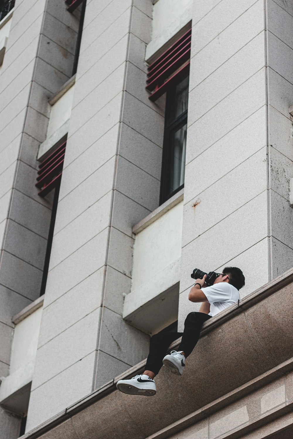 白いTシャツと黒いサングラスをかけた男が、昼間、茶色のコンクリートの壁に座っている