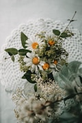 A bouquet featuring white daisies with yellow centers is arranged against a delicate crocheted doily background. The daisies are complemented by small green leaves and clusters of tiny wildflowers. The composition gives a sense of natural elegance and simplicity.