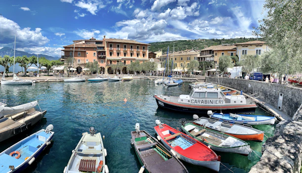 A picturesque marina scene featuring a collection of small boats docked along clear, turquoise waters. A Carabinieri patrol boat is prominently visible among other colorful boats. The background displays charming, multi-story buildings with arches, surrounded by lush greenery and olive trees, under a partly cloudy sky.