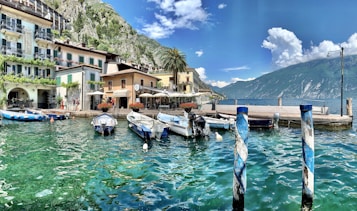 A picturesque waterfront scene with boats moored along a vibrant turquoise canal. Colorful buildings lined with balconies and ornate architecture sit against a lush green mountainside. Palm trees, cafes with outdoor seating, and a mountainous backdrop contribute to the scenic beauty. Fluffy white clouds dot the clear blue sky.