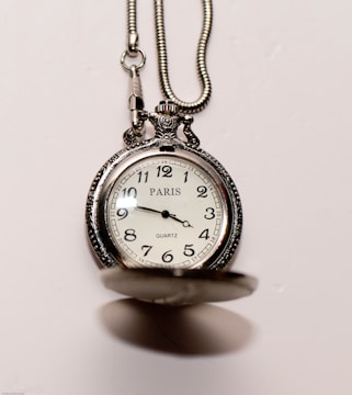 An ornate pocket watch with a silver chain is featured. The clock face displays the time and has markings for each hour, with the word 'PARIS' prominently displayed. The cover of the pocket watch is slightly open, revealing the classic watch face.