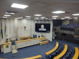 A large, empty municipal chamber with rows of blue seats arranged in a semi-circular fashion around a central podium. The walls are made of light-colored stone with flags and a crucifix adorning one wall. A large digital screen displays the emblem and name of São Paulo's city council. The atmosphere is quiet and formal, suitable for official meetings.