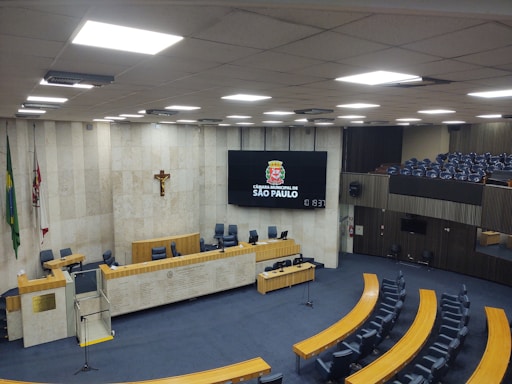 A large, empty municipal chamber with rows of blue seats arranged in a semi-circular fashion around a central podium. The walls are made of light-colored stone with flags and a crucifix adorning one wall. A large digital screen displays the emblem and name of São Paulo's city council. The atmosphere is quiet and formal, suitable for official meetings.