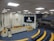 A large, empty municipal chamber with rows of blue seats arranged in a semi-circular fashion around a central podium. The walls are made of light-colored stone with flags and a crucifix adorning one wall. A large digital screen displays the emblem and name of São Paulo's city council. The atmosphere is quiet and formal, suitable for official meetings.