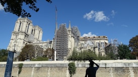 A historic Gothic cathedral undergoing restoration, with scaffolding covering sections of the facade. Trees line the front while a person in silhouette stands in the foreground. The sky is clear with a few scattered clouds.