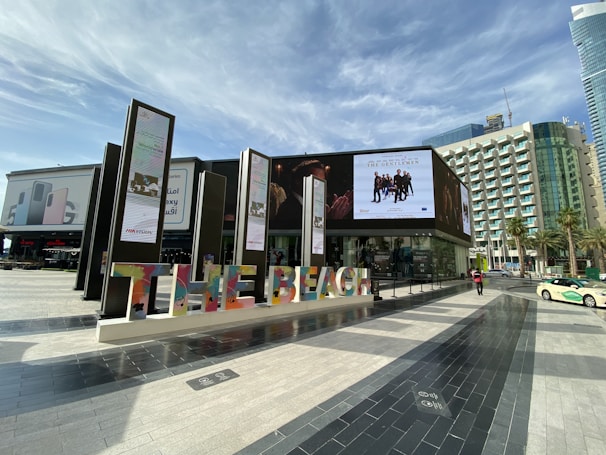 An outdoor shopping and entertainment area featuring modern architecture with a large digital screen displaying an advertisement for 'The Gentlemen' movie. Colorful letters spelling 'THE BEACH' are prominently displayed. Surrounding buildings include a skyscraper and other commercial structures. The area is paved with decorative tiles and is set under a partly cloudy sky.