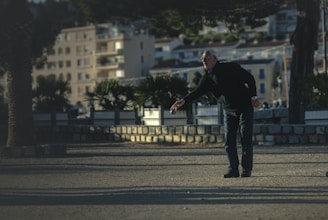 An older man wearing dark clothing is playing a game of pétanque in an open outdoor space surrounded by trees and residential buildings. He appears to be focused as he throws a metal ball, with sunlight casting long shadows on the ground.